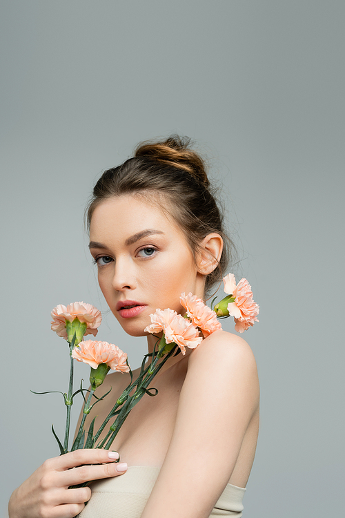 pretty young woman with natural makeup and bare shoulders looking at camera near peach carnations isolated on grey