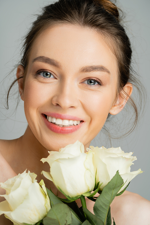 Portrait of cheerful young woman looking at camera near rose flowers isolated on grey