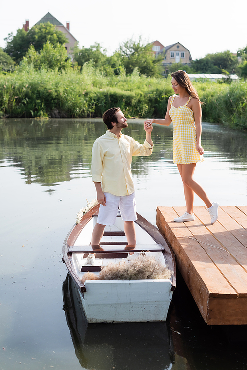 side view of happy bearded man in summer clothes holding hands with cheerful girlfriend near boat