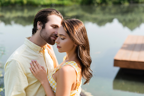 young woman with closed eyes hugging with bearded man near lake