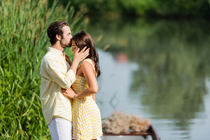 side view of bearded man kissing forehead of young woman in dress near lake