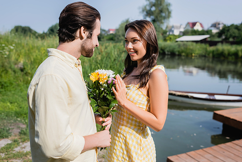 bearded man holding bouquet of flowers near happy woman in dress near lake