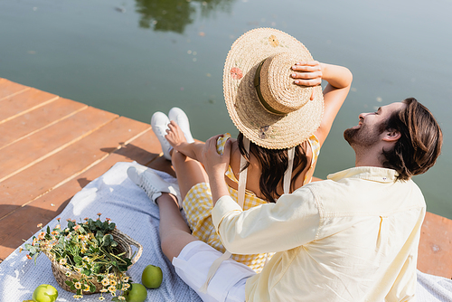 high angle view of cheerful man looking at girlfriend in straw hat while sitting on pier during picnic