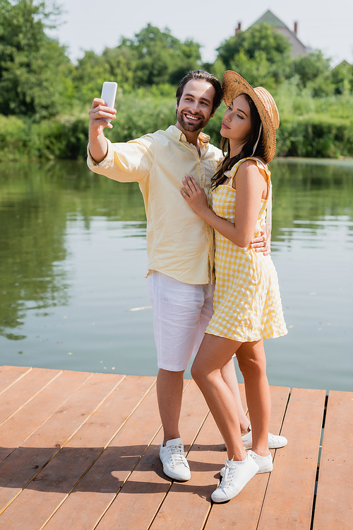 full length of happy young couple taking selfie near lake