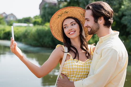 happy young woman in straw hat taking selfie with boyfriend near lake
