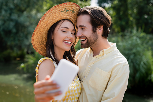 happy young woman in straw hat taking selfie with joyful boyfriend outdoors