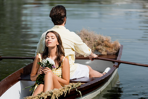 young woman with closed eyes holding flowers and leaning on back of boyfriend during romantic boat trip
