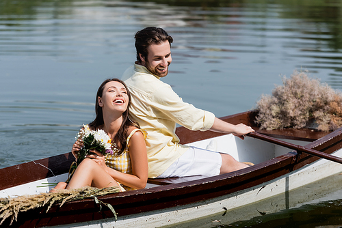 happy young woman holding flowers and leaning on back of boyfriend during romantic boat trip