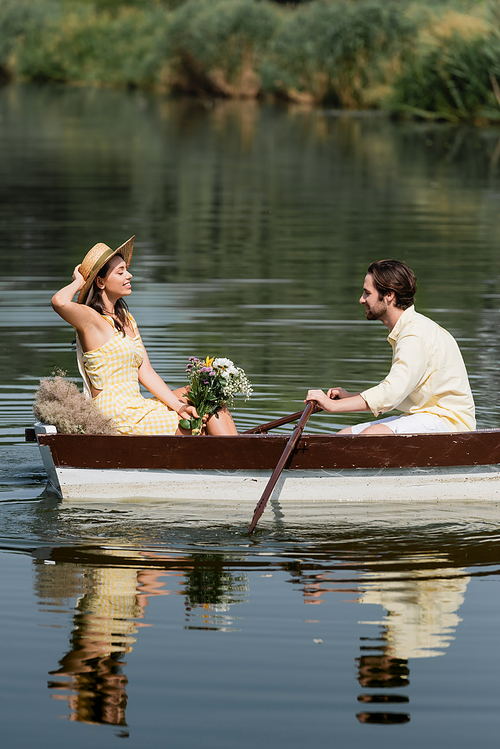 side view of smiling woman in straw hat holding flowers and having romantic boat ride with man