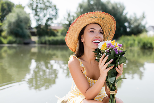joyful young woman in straw hat and dress holding flowers near lake