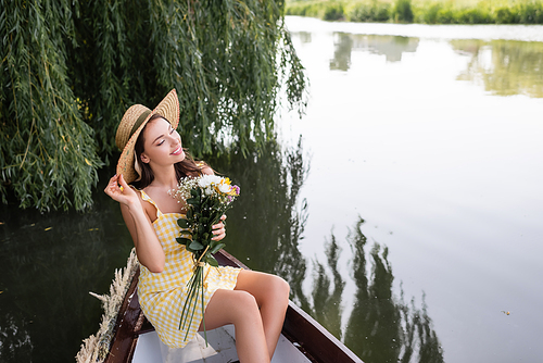 dreamy young woman adjusting straw hat and holding flowers during boat ride on lake