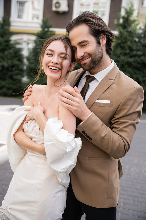 portrait of positive and bearded man hugging happy bride in wedding dress