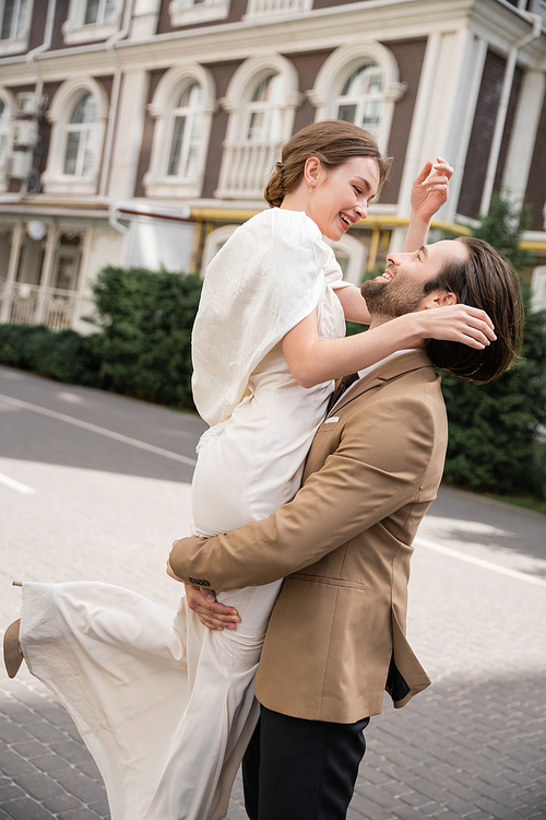 bearded groom in formal wear lifting happy bride in white dress outside