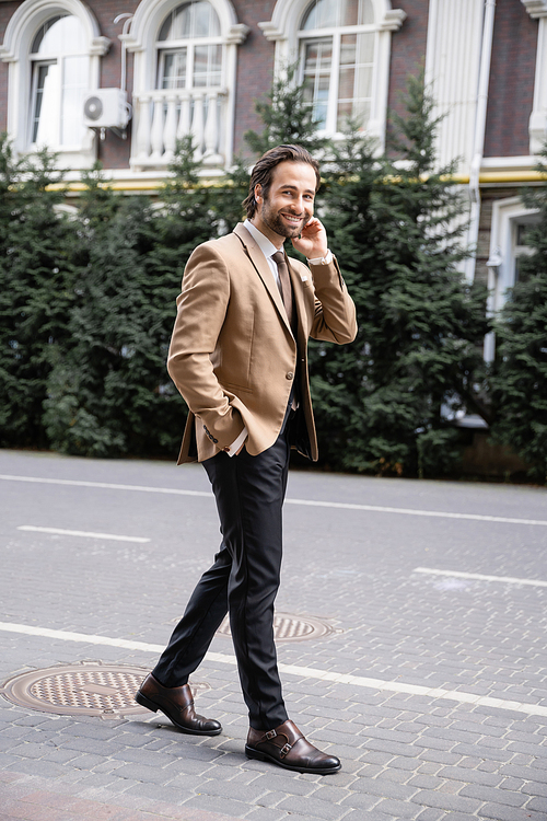 full length of cheerful man in beige formal wear standing with hand in pocket and looking at camera