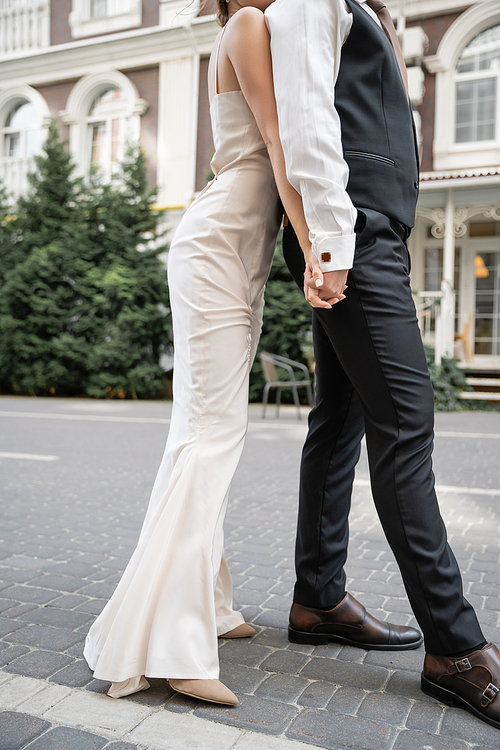 cropped view of young bride and groom in wedding dress and suit holding hands outside