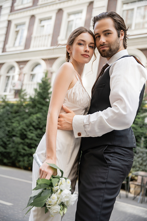 low angle view of bearded groom in vest embracing bride in white dress with wedding bouquet