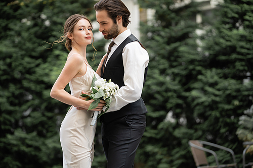 bearded groom in vest embracing bride in wedding dress with bouquet