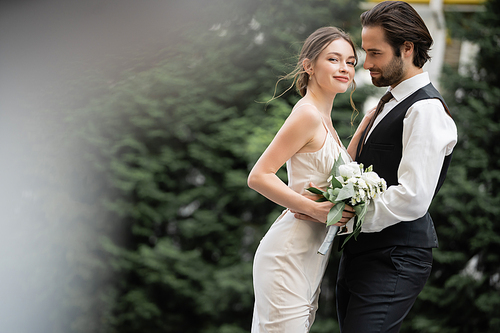 bearded groom in vest embracing happy bride in white dress with wedding bouquet