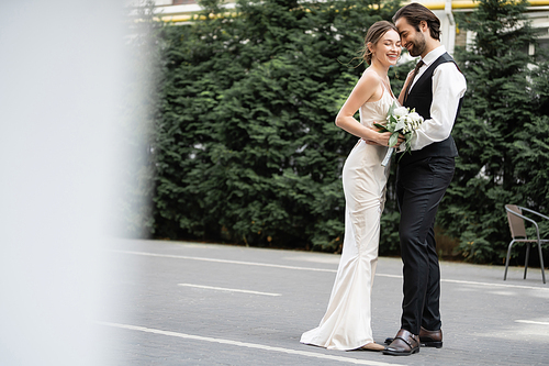 happy groom in vest embracing cheerful bride in white dress with wedding bouquet