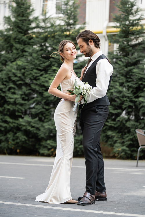 full length of bearded groom in vest embracing cheerful bride in white dress with wedding bouquet