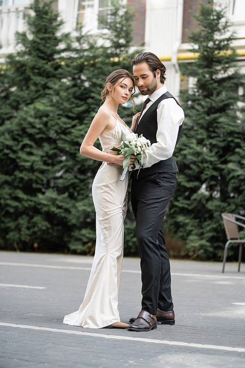 full length of bearded man in vest embracing bride in white dress with wedding bouquet