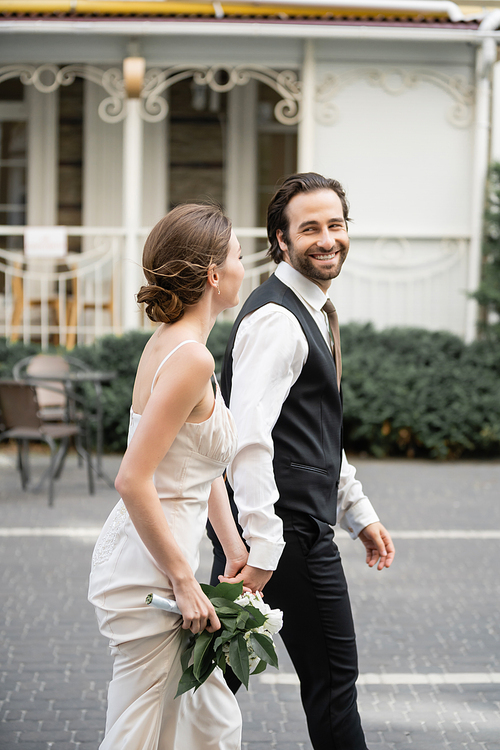 happy groom holding hand of bride in white dress with bouquet of flowers