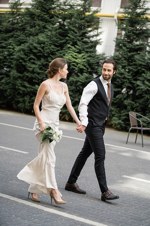 full length of groom holding hand of bride in white dress with bouquet of flowers