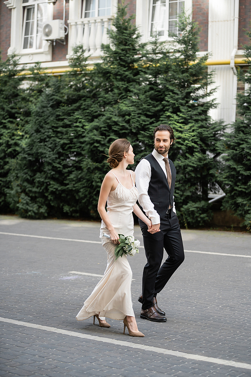 full length of man in suit holding hand of bride in white dress with bouquet of flowers