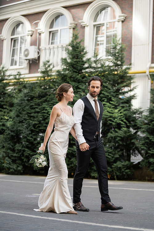 full length of groom walking with happy bride in white dress holding bouquet of flowers