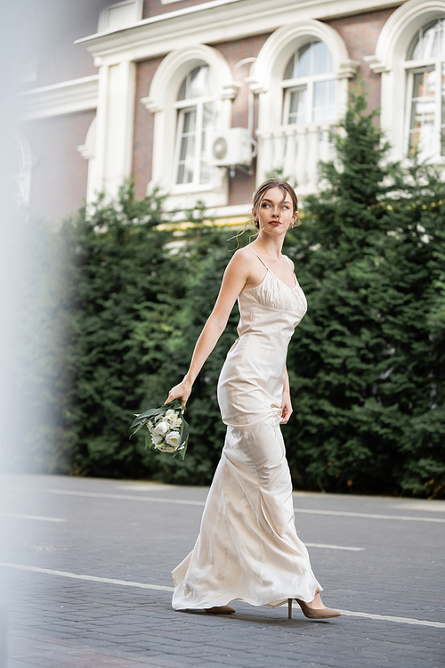 full length of young bride in white dress holding wedding bouquet of blooming flowers and walking outside