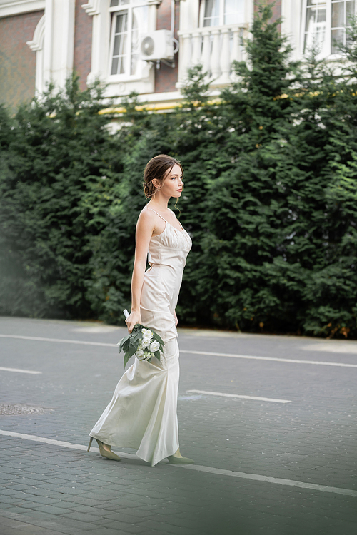 full length of young woman in white dress holding wedding bouquet of flowers and standing outside