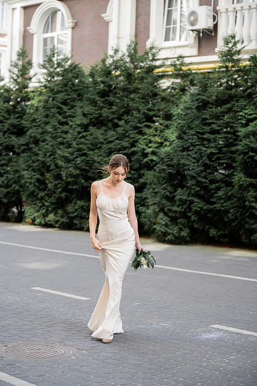 full length of young bride in white dress holding wedding bouquet of flowers and walking on street