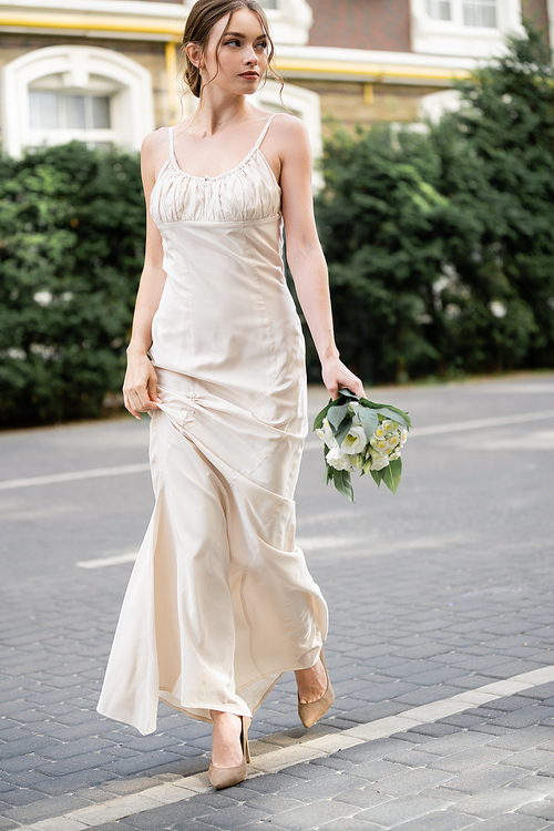 full length of young bride in white dress holding wedding bouquet with flowers