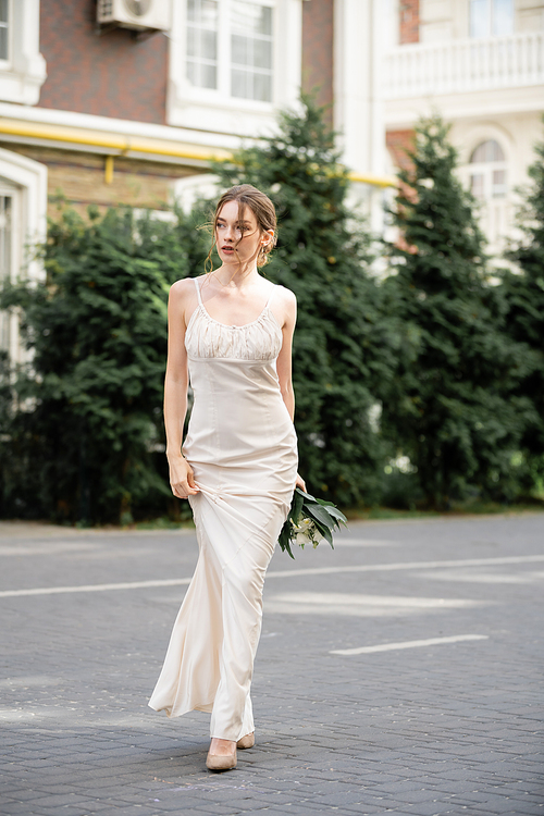 full length of pretty bride in white dress holding wedding bouquet and walking on street