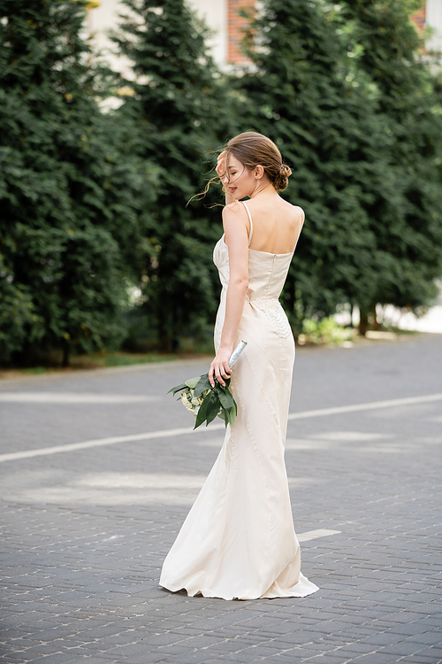 full length of happy bride in white dress holding wedding bouquet and standing on street