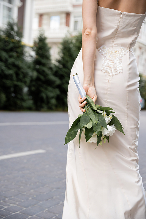 cropped view of young woman in white dress holding wedding bouquet behind back