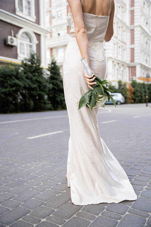 partial view of young woman in white dress holding wedding bouquet behind back