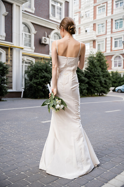 back view of young woman in white dress holding wedding bouquet behind back