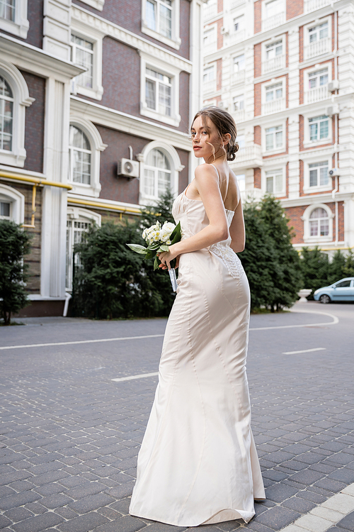 full length view of young woman in white dress holding wedding bouquet