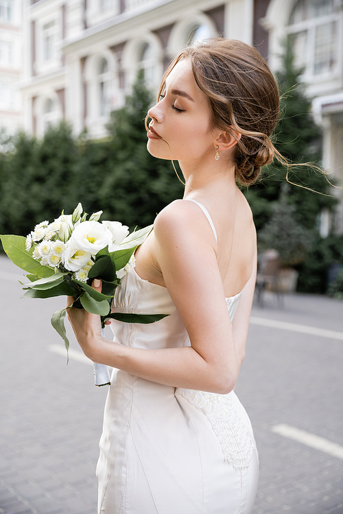 young bride in white dress standing with closed eyes and holding wedding bouquet