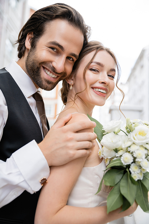 portrait of happy groom with beard hugging cheerful bride holding wedding bouquet