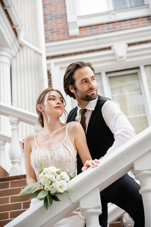 young woman in wedding dress and groom in suit looking away near house