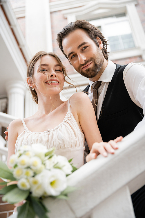 low angle view of happy young woman in wedding dress and groom in suit looking at camera near house