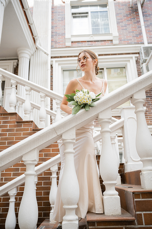 bride in wedding dress holding bouquet and looking away near house