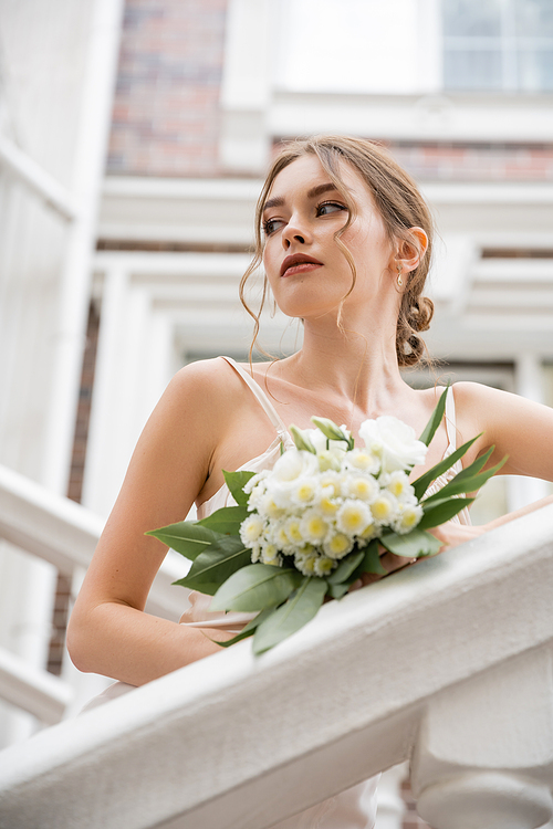 low angle view of bride in wedding dress holding bouquet and looking away near house