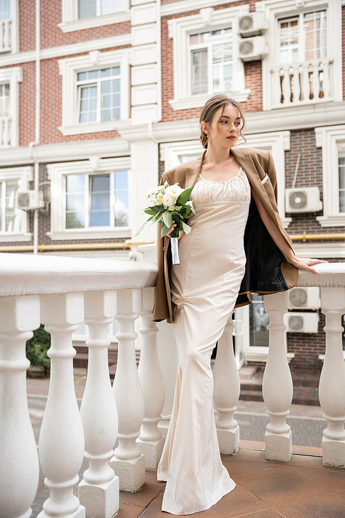 full length of young bride in wedding dress and beige blazer holding bouquet near house