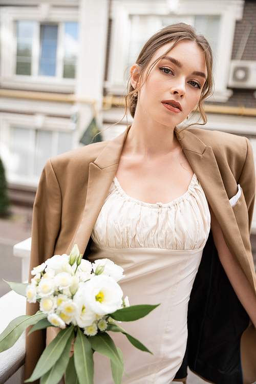 young bride in wedding dress and beige blazer holding bouquet and looking at camera