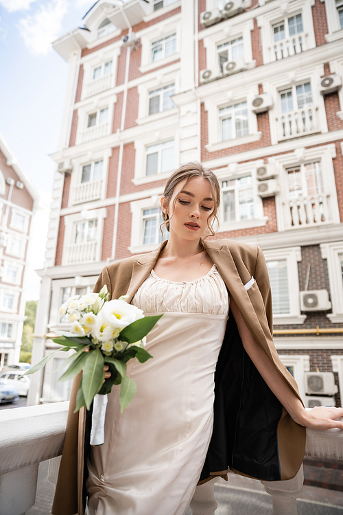 young woman in wedding dress and beige blazer holding bouquet of flowers