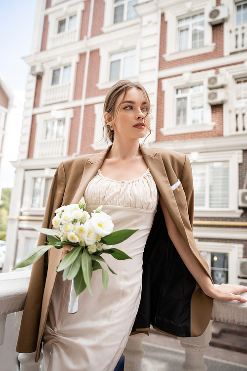 pretty young woman in wedding dress and beige blazer holding bouquet of flowers