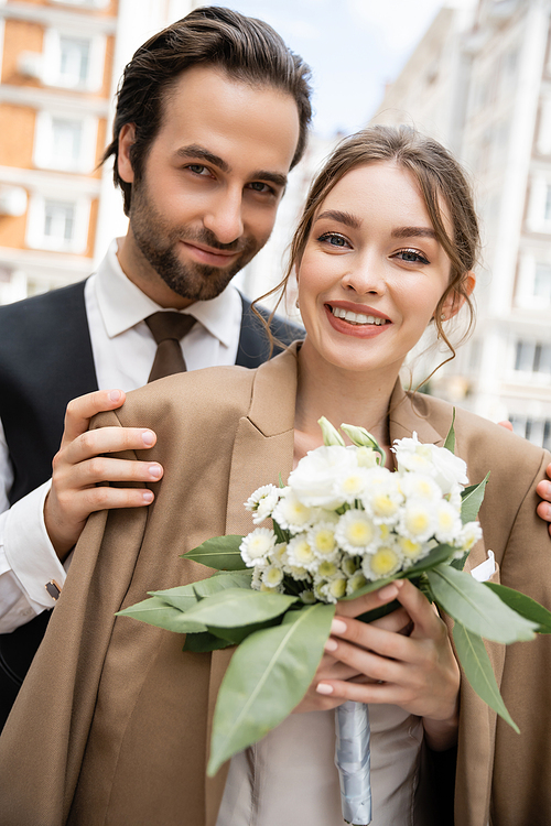 portrait of happy groom in vest hugging cheerful bride in beige blazer and holding wedding bouquet